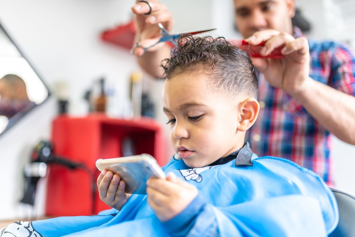 Cute Little Boy Getting a Hair Cut in a Barber Shop or Salon. Holding a Cell Phone While Getting a Hair Cut. Beauty Concept.